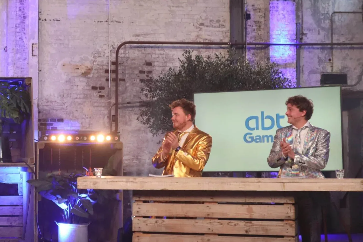 Kenneth en Andor in gouden en zilveren pakken achter de quiztafel tijdens een bedrijfsshow