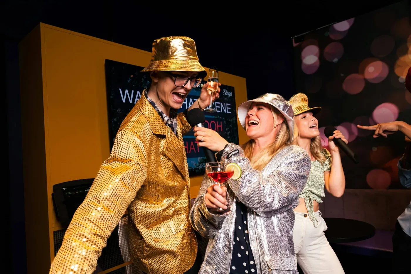 Twee gasten in glitteroutfits zingen samen in een microfoon